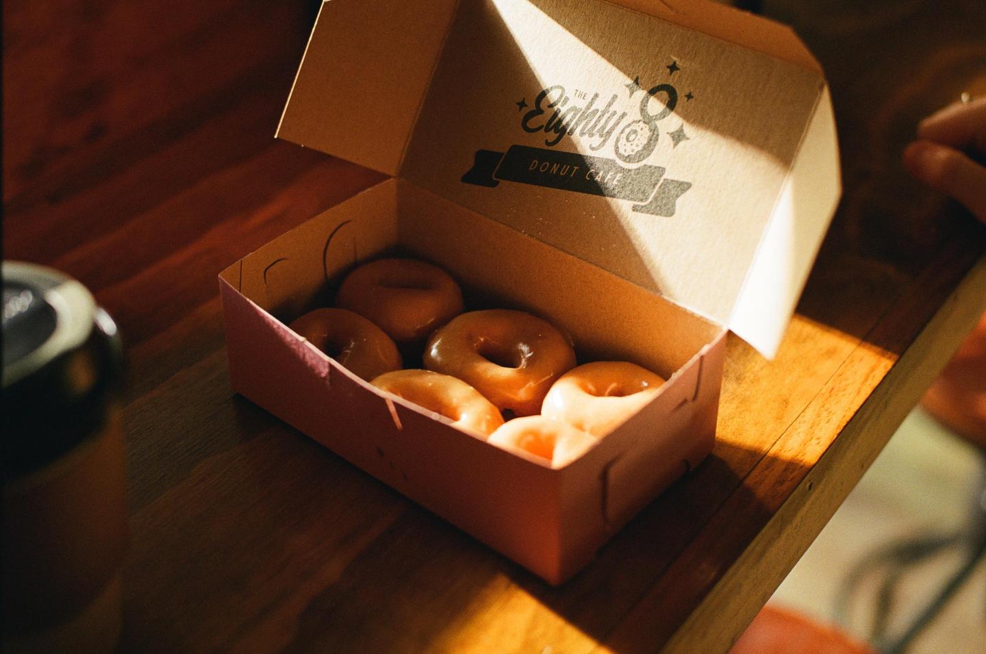 A box of glazed donuts sits on a wooden surface, illuminated by a shaft of sunlight.