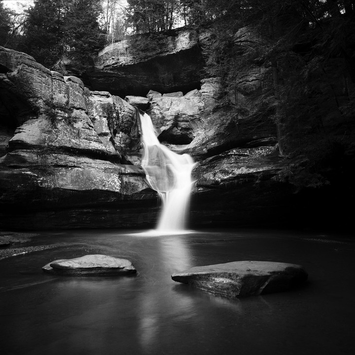 A waterfall cascades into a pool with two rocks in the foreground, surrounded by rocky cliffs and trees.