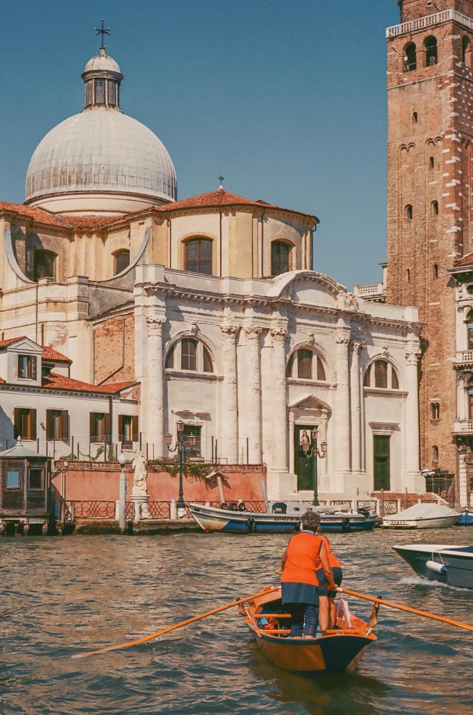 A person rows a boat on a canal in Venice with historic buildings in the background.