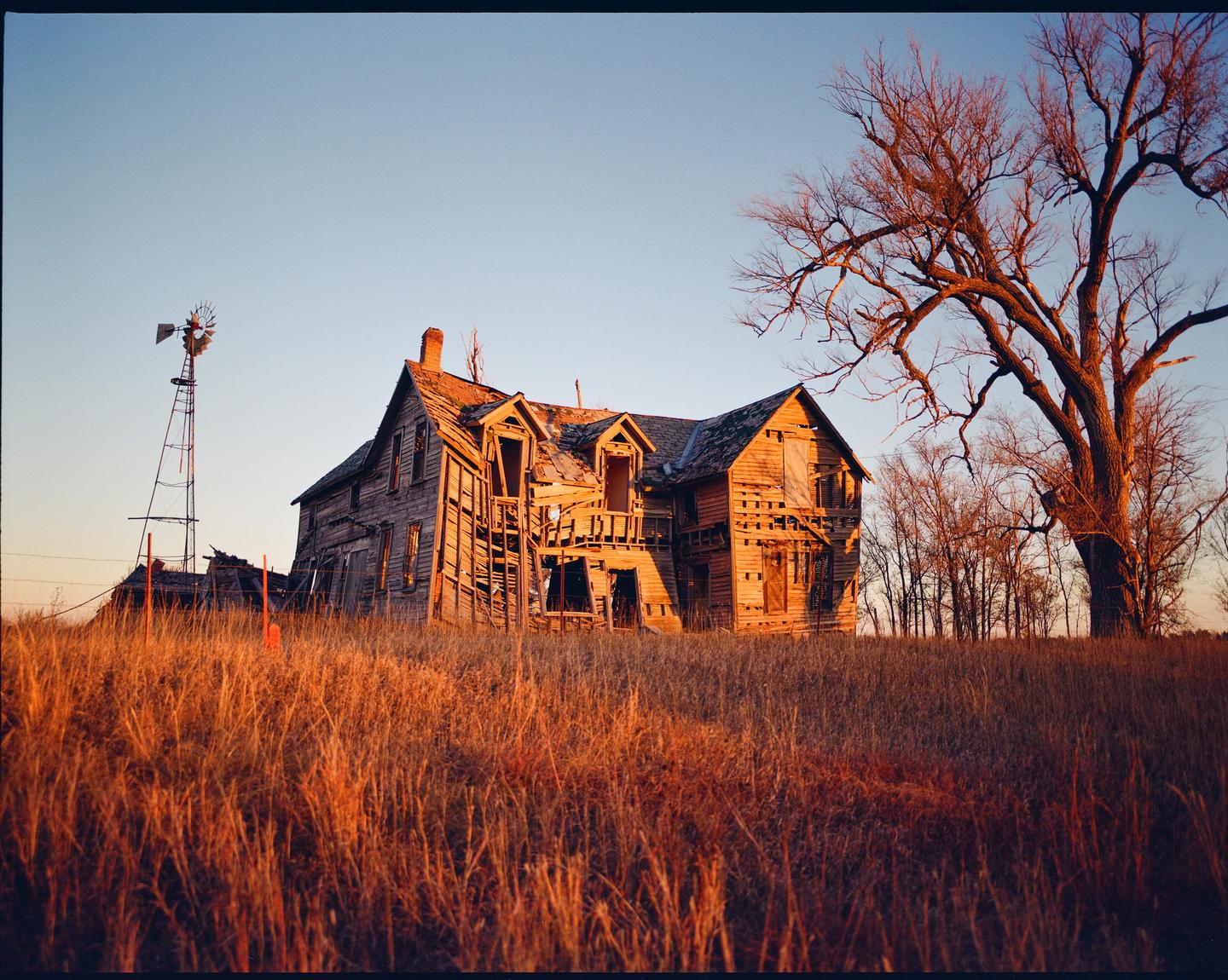 An abandoned wooden house stands in a field of tall grass with a windmill and a large bare tree under a clear sky.