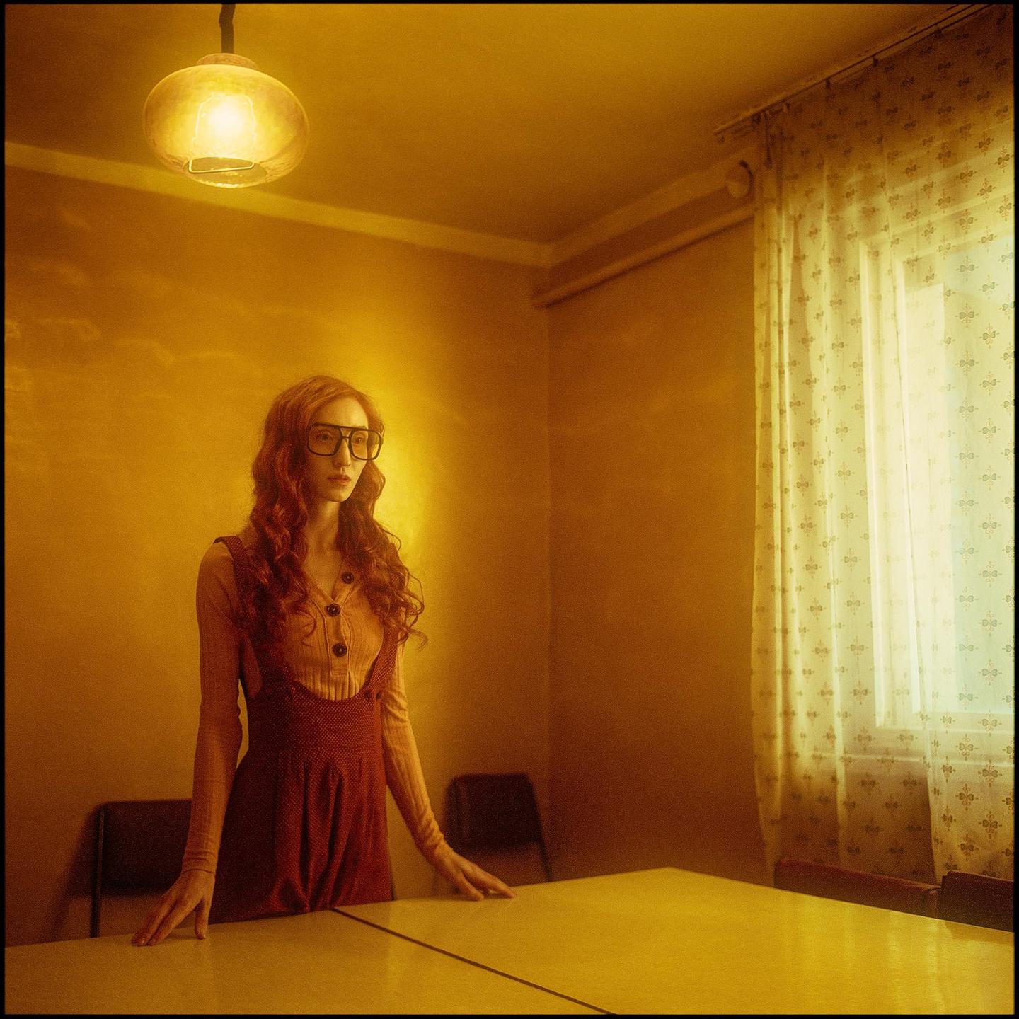 A woman with long curly hair and glasses stands at a table in a room with a window and a hanging lamp.