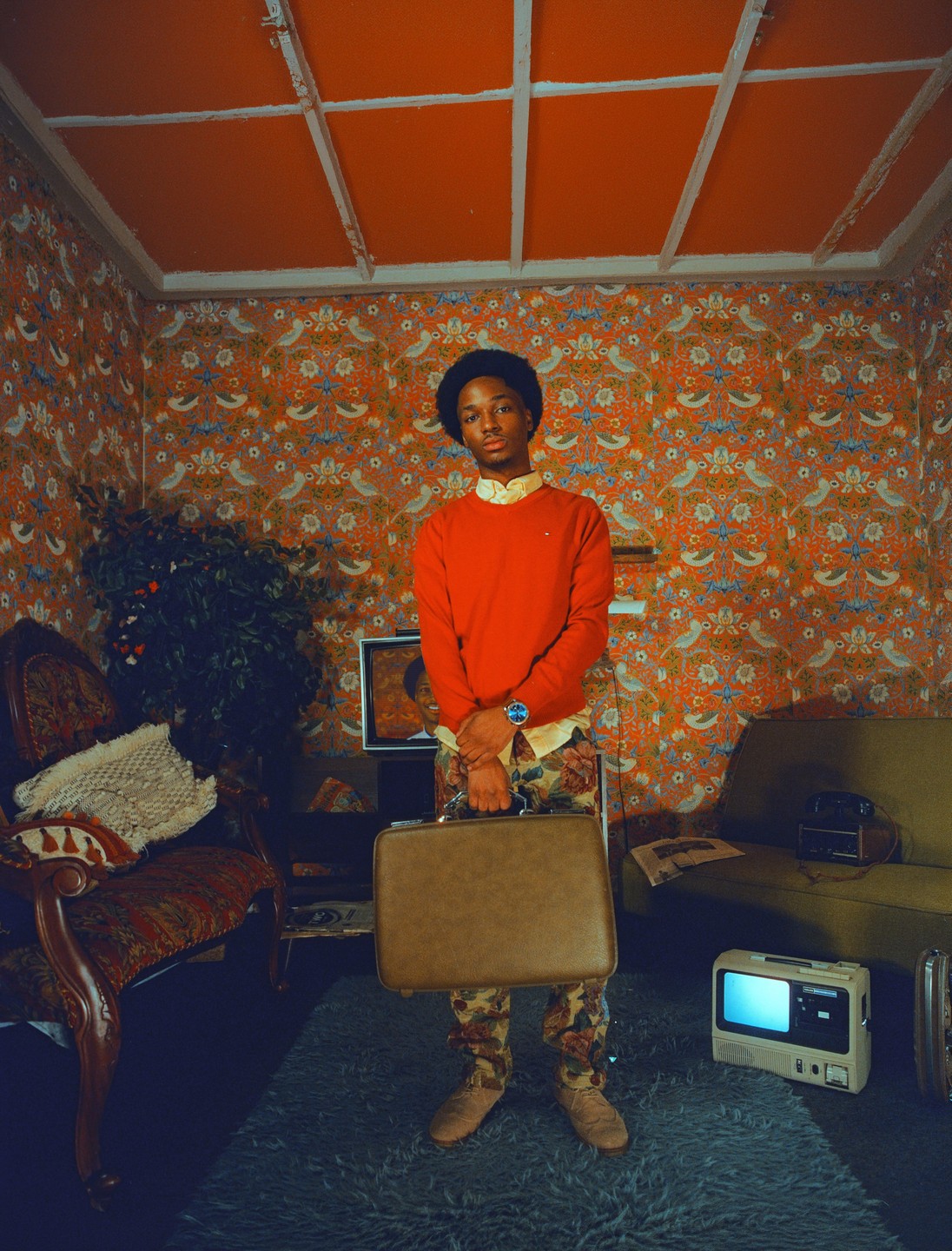 A man stands in a room with patterned wallpaper, holding a briefcase.