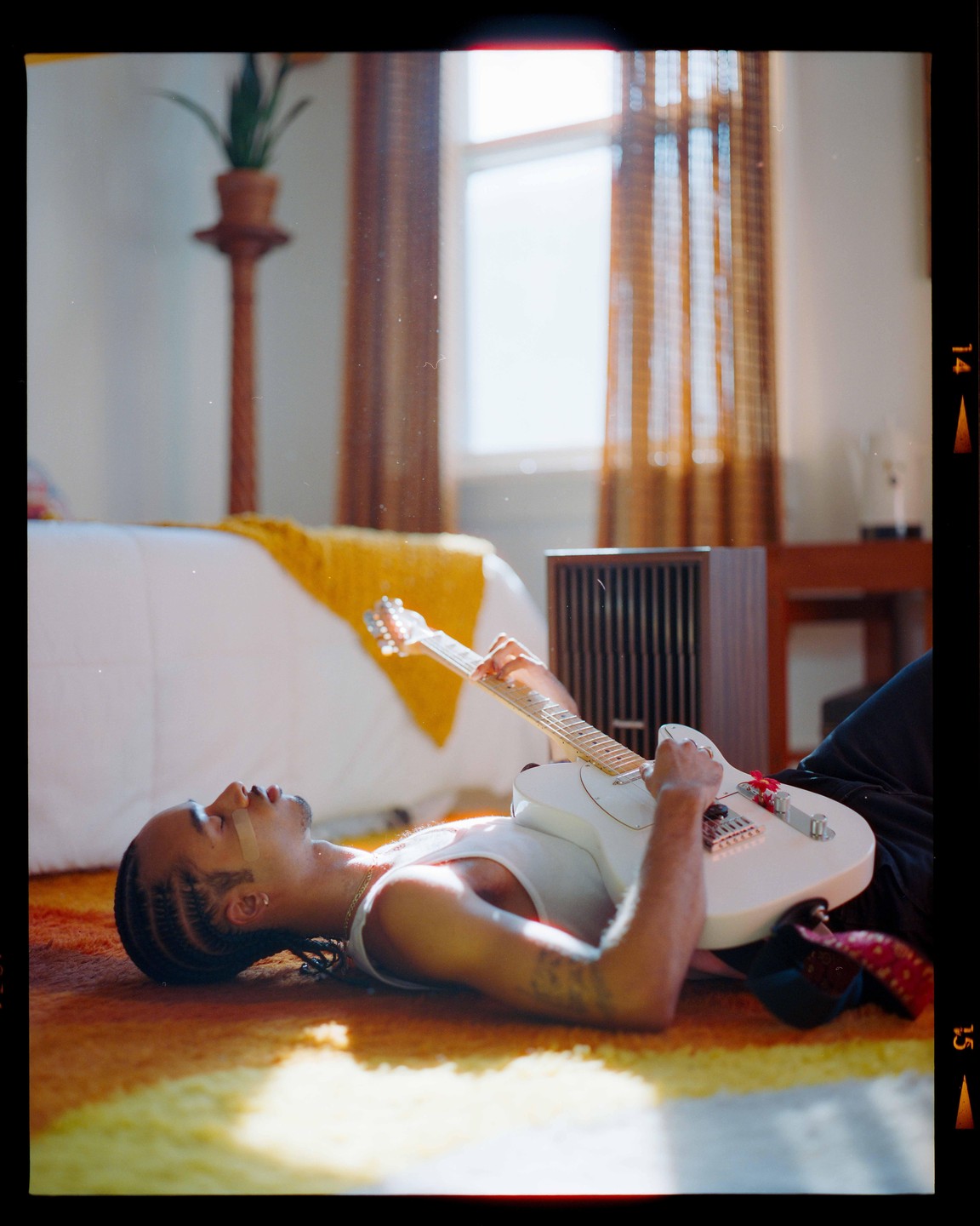 A man lies on a colorful rug, playing an electric guitar in a sunlit room.