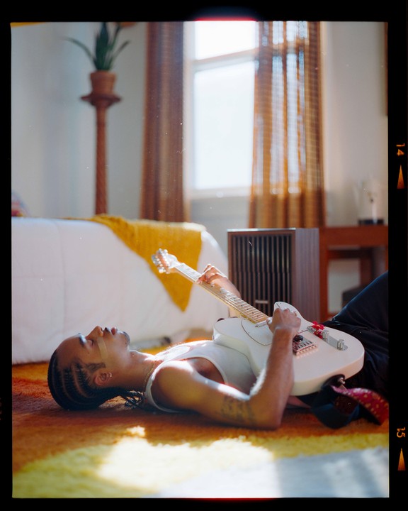 A man lies on a colorful rug, playing an electric guitar in a sunlit room.