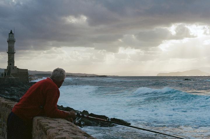 A man in a red sweater fishes from a stone wall as waves crash against the shore near a lighthouse under a cloudy sky.