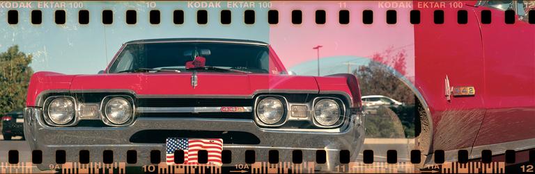 A red vintage car with a chrome bumper and an American flag license plate is parked outdoors.