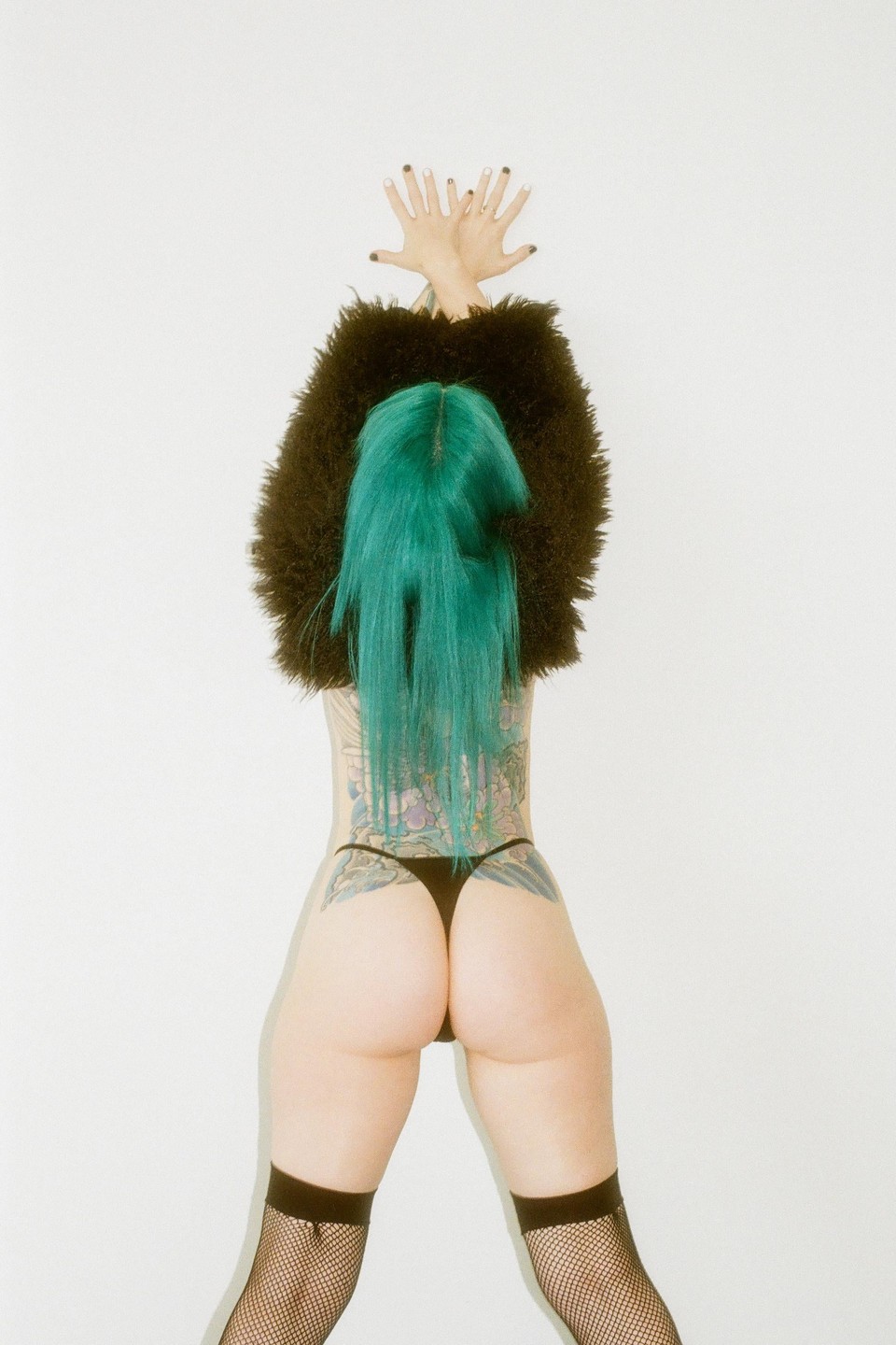 A woman with green hair and a black furry top stands with her back to the viewer, hands raised above her head.