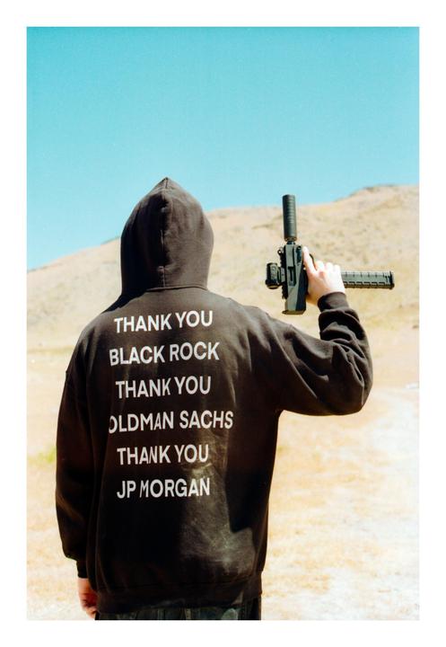 A person wearing a black hoodie with white text on the back holds a firearm in a desert landscape.