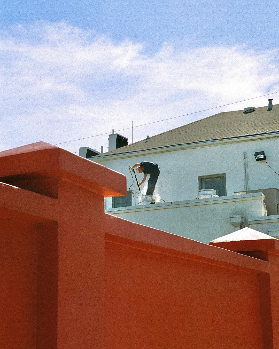 A man pressure washes a roof behind a bright orange wall under a blue sky.