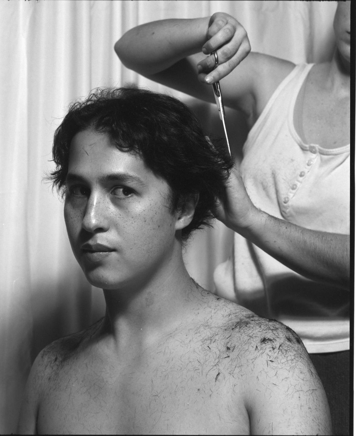A man with short hair looks at the camera while another person cuts his hair, with hair clippings visible on his bare shoulders.