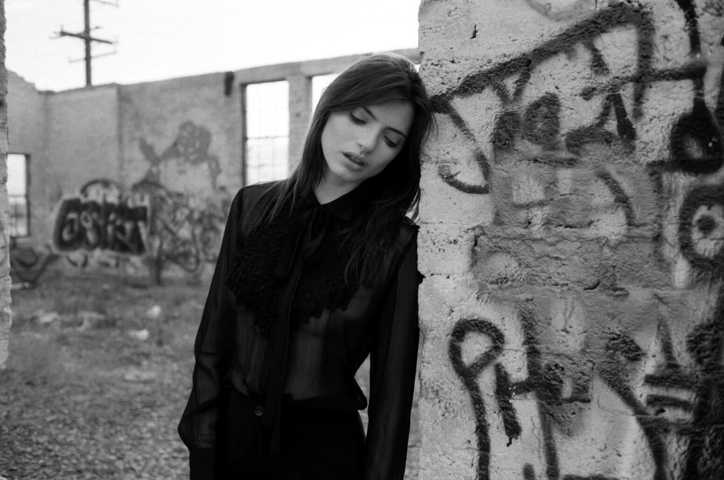 A woman with long hair leans against a graffiti-covered wall in a ruined building.