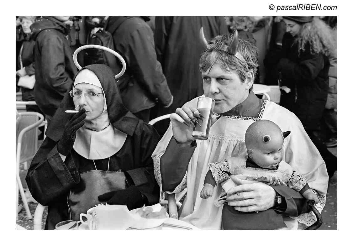 A woman dressed as a nun with a halo smokes a cigarette next to a man dressed as a devil holding a doll with horns and a third eye.