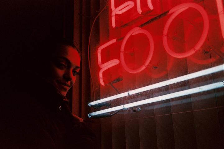 A woman stands next to a red neon sign that reads "FOO" and two fluorescent tubes.