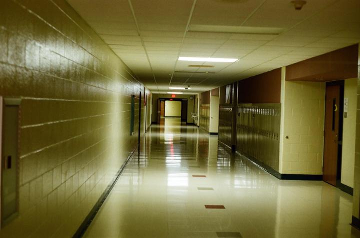 A long, empty school hallway stretches into the distance, lined with lockers and illuminated by overhead lights.