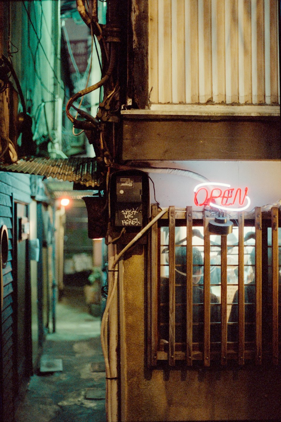 A neon "OPEN" sign glows above a window with wooden bars, next to a narrow alleyway at night.