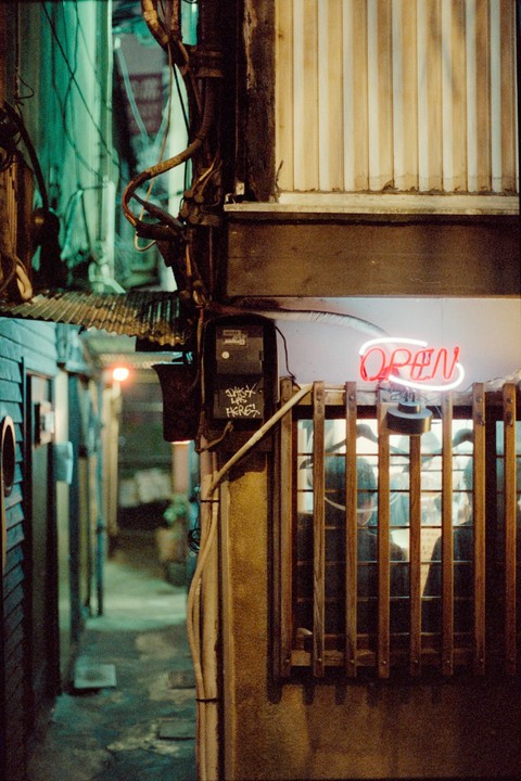 A neon "OPEN" sign glows above a window with wooden bars, next to a narrow alleyway at night.