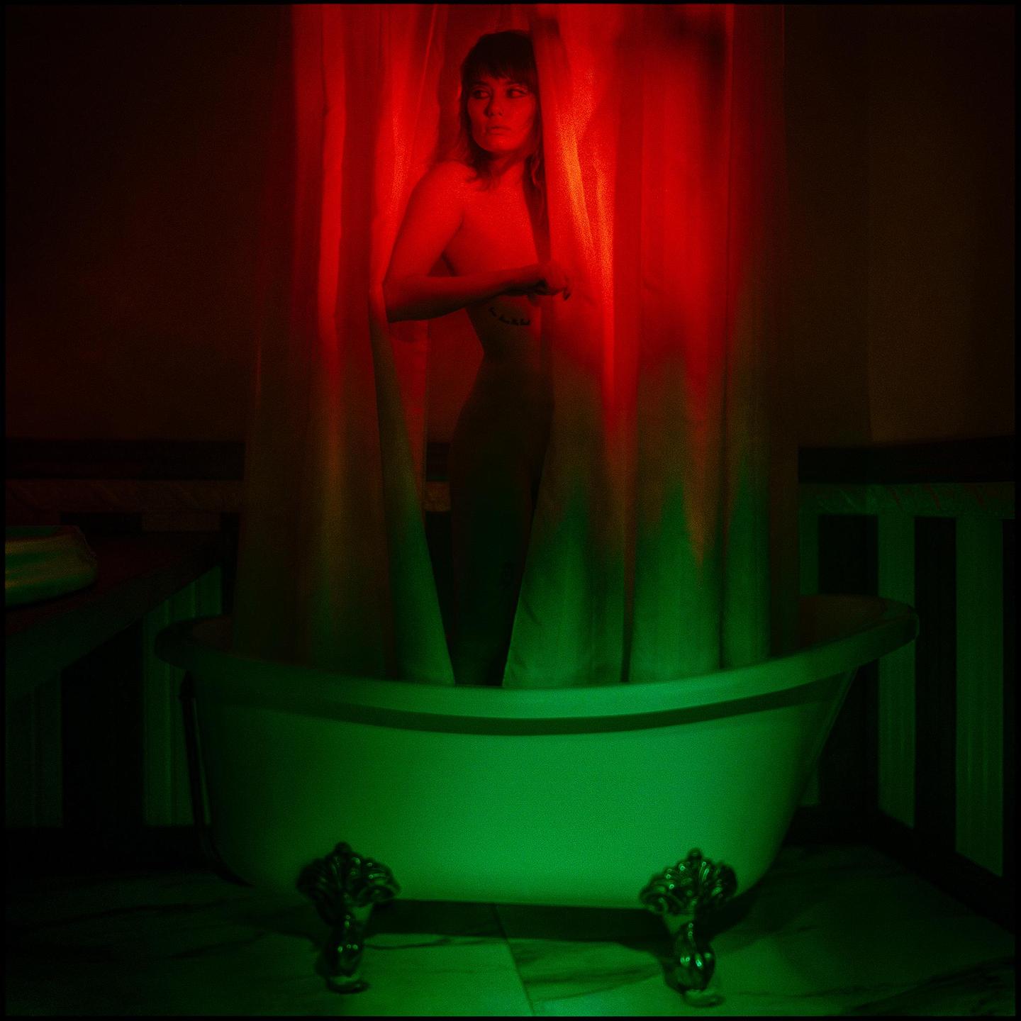 A woman stands behind a shower curtain in a clawfoot bathtub, illuminated by red and green light.