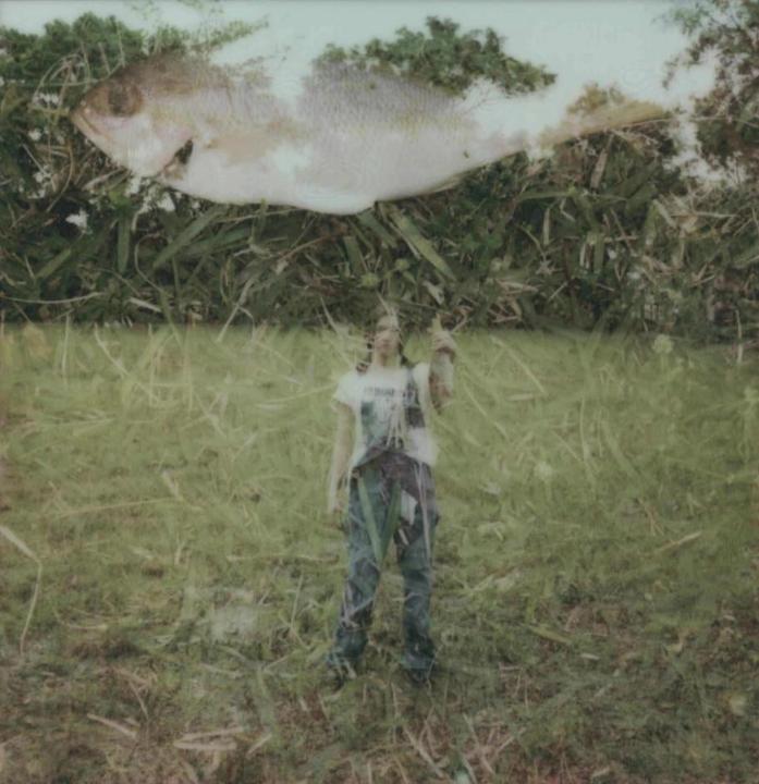 A person stands in a grassy field holding a small fish, with a large fish superimposed above them.