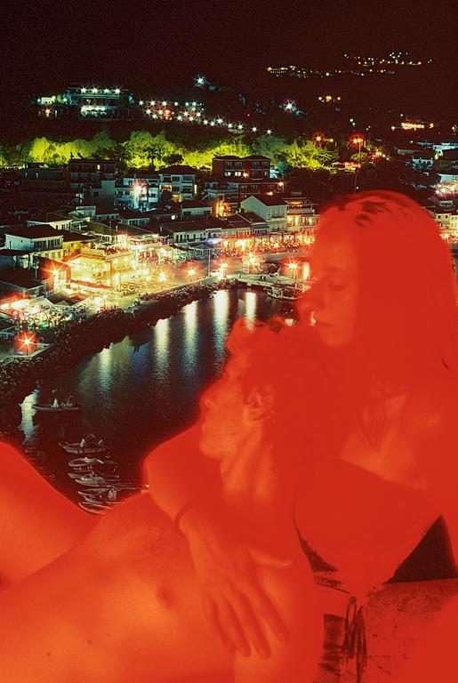 A couple is embracing in the foreground with a brightly lit town and harbor visible in the background at night.