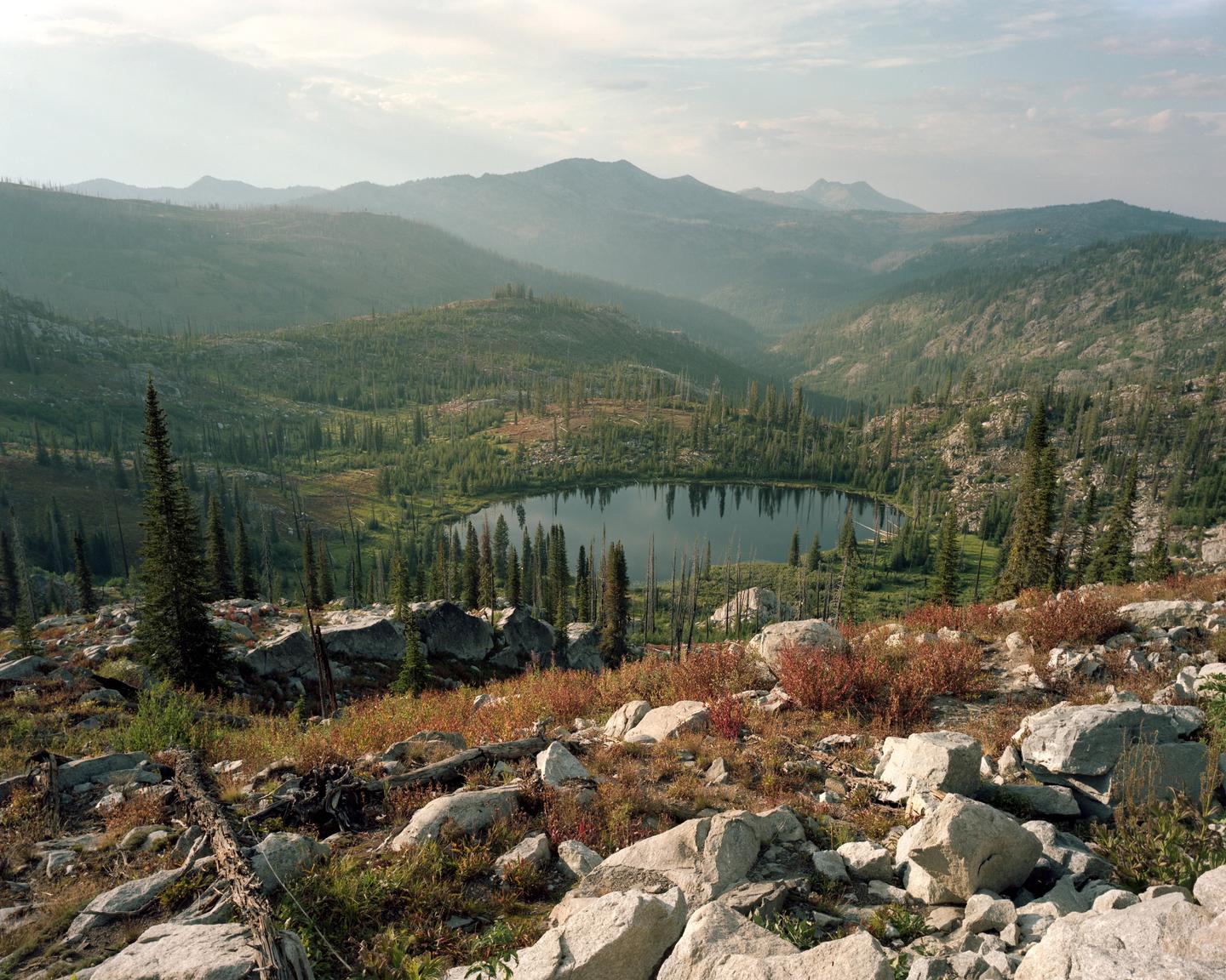 A mountain lake is surrounded by a forest of pine trees and rocky terrain under a hazy sky.