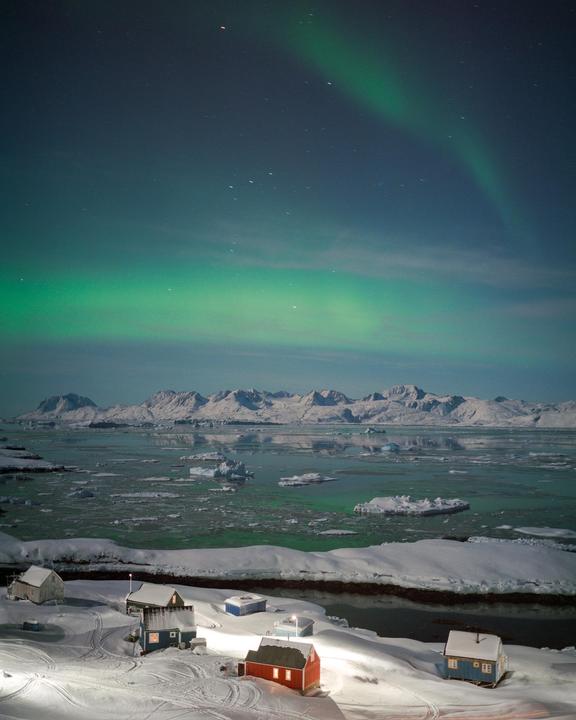 A small village with colorful houses sits on a snow-covered shore under a vibrant aurora borealis and a starry night sky, with mountains and ice-filled water in the distance.
