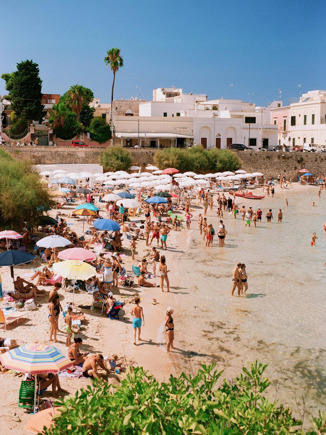 A crowded beach is filled with people swimming and sunbathing under colorful umbrellas, with white buildings and palm trees in the background.