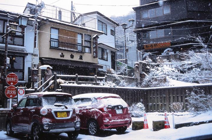 A snowy street scene in Japan shows two red cars parked in front of buildings, with a person holding an umbrella walking on a raised pathway.