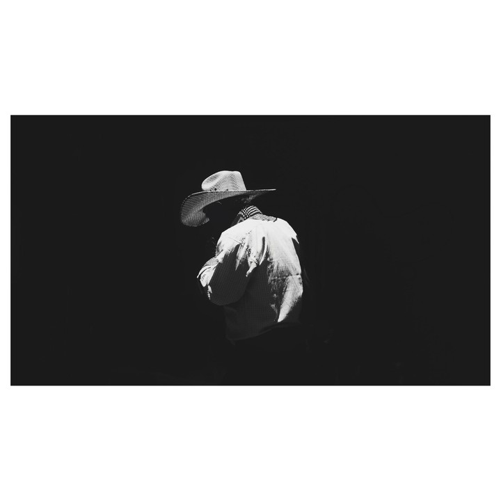 A man in a cowboy hat is illuminated by sunlight from behind against a dark background.