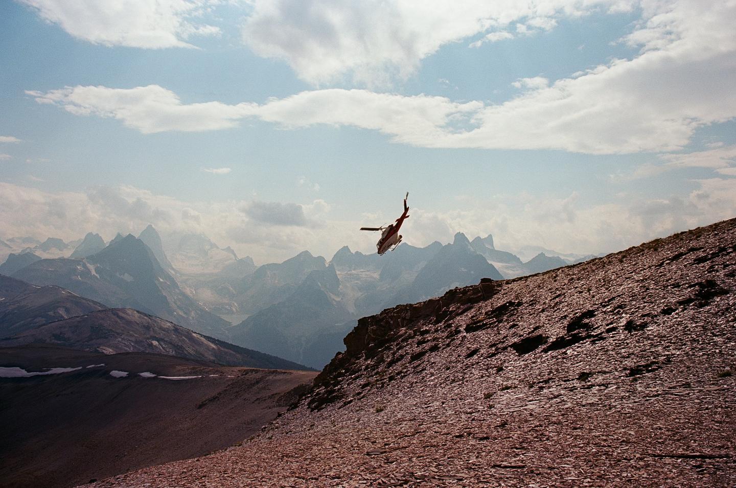 A red and white helicopter flies over a rocky mountain slope with a range of mountains in the background under a cloudy sky.