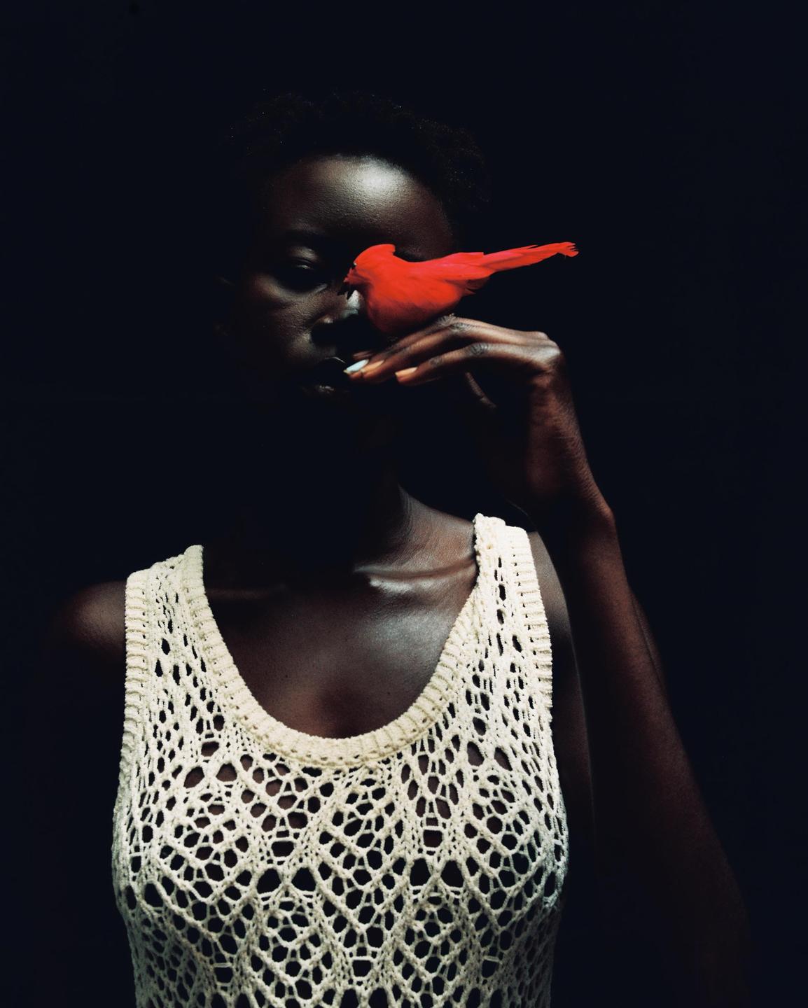 A woman in a white crocheted top holds a red bird to her face.