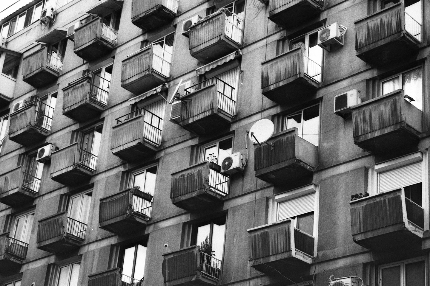 A black and white photograph captures the facade of a building adorned with numerous balconies, some featuring satellite dishes and air conditioning units.