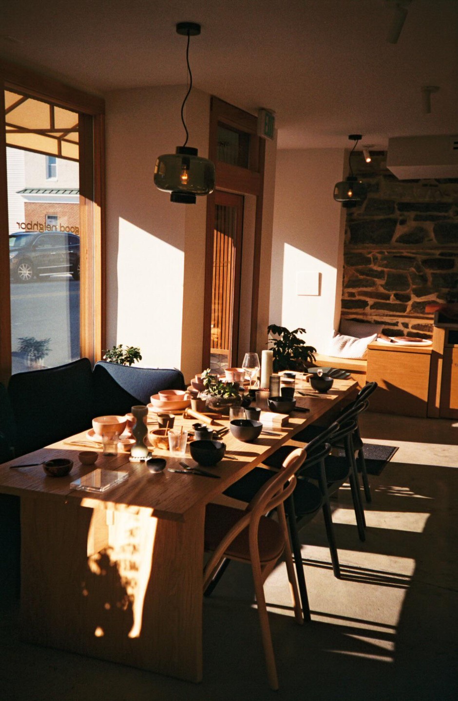 A long wooden table is set with various bowls and glasses in a sunlit room with a stone wall.