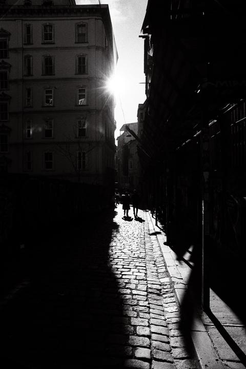 Two people walk on a sunlit cobblestone street between tall buildings, with one side of the street in deep shadow.