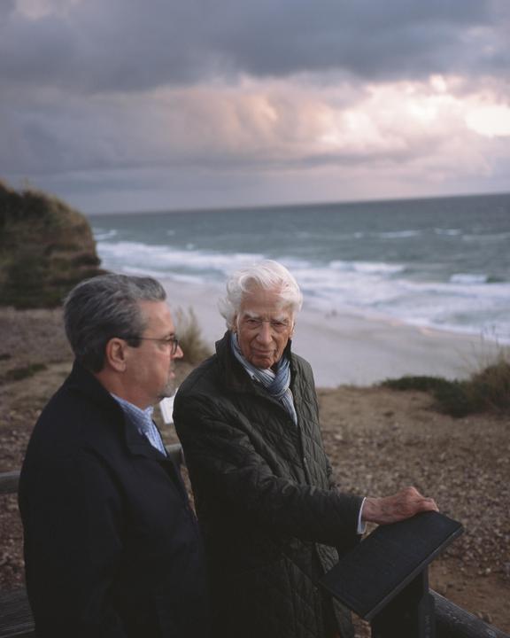 Two men stand on a cliff overlooking a beach and ocean under a cloudy sky.