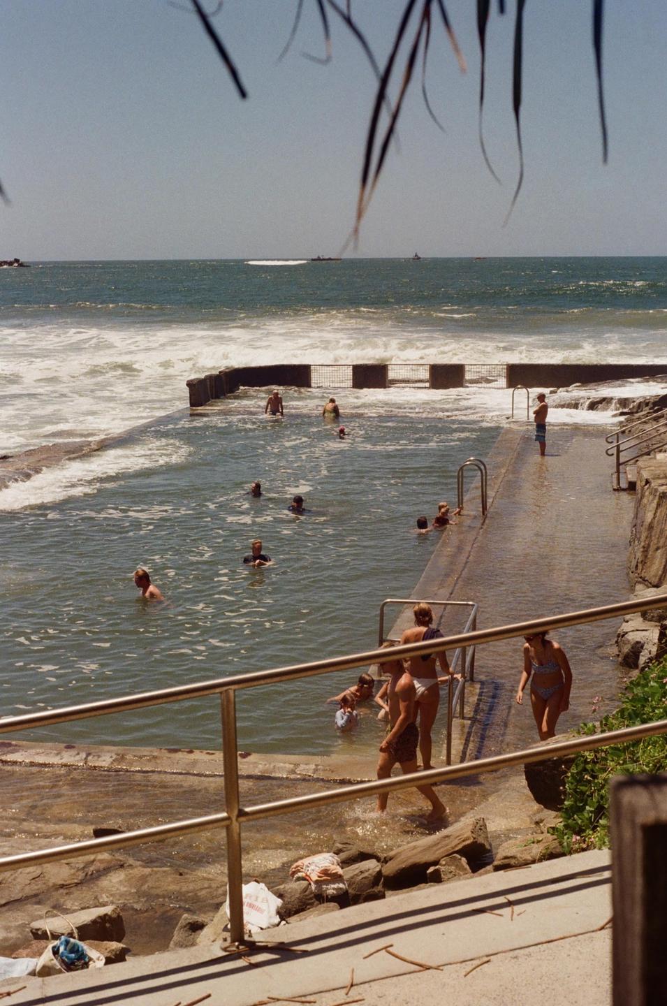 People swim in a concrete ocean pool next to a rocky shore with waves crashing over the wall.