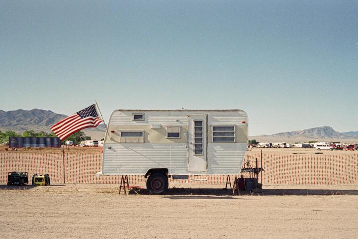 An American flag flies next to a white travel trailer in a desert landscape with mountains in the distance.