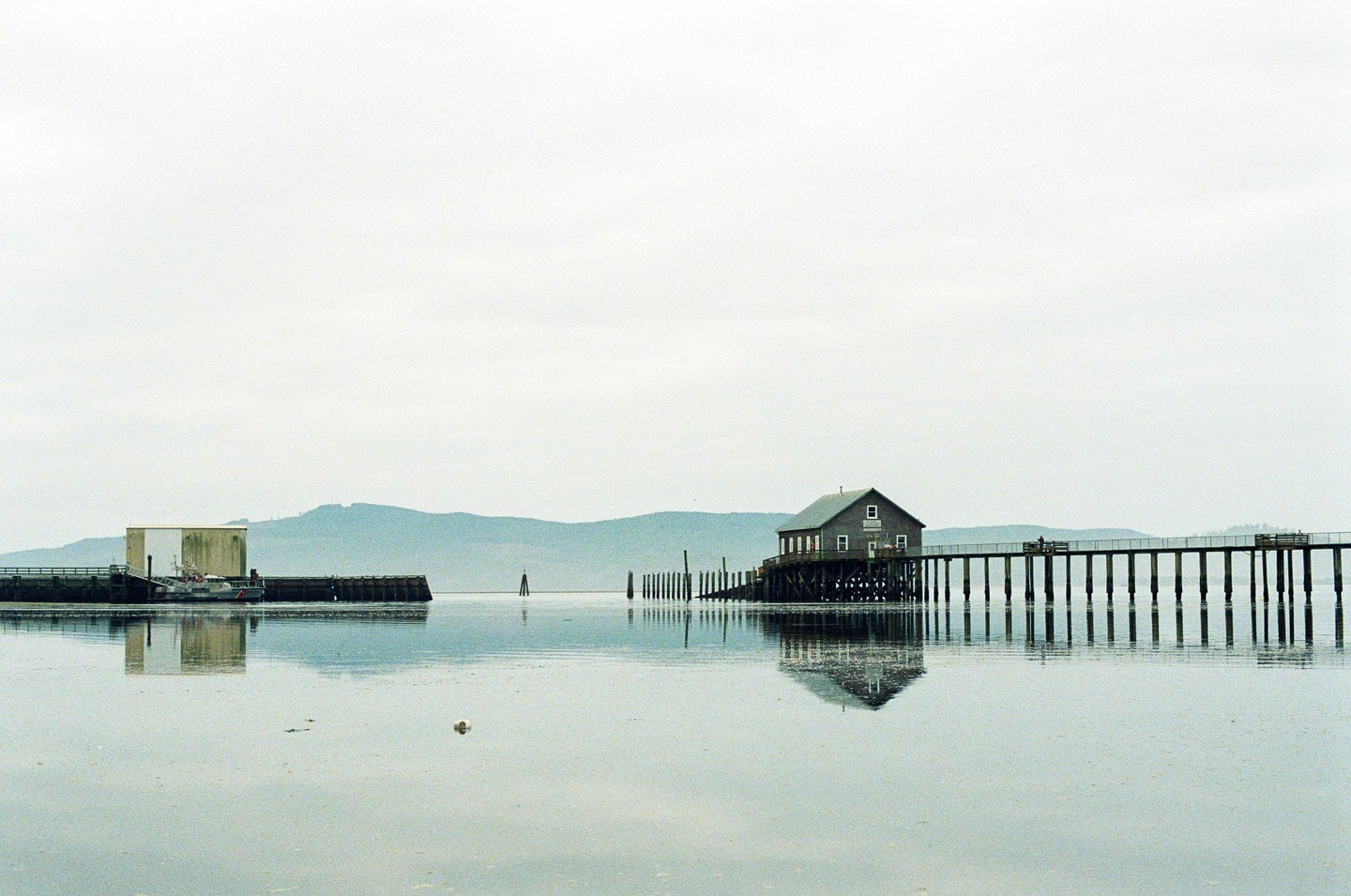 A house on stilts is connected to a pier over calm water with mountains in the background.