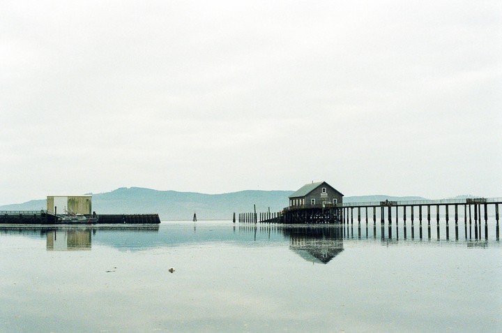 A house on stilts is connected to a pier over calm water with mountains in the background.