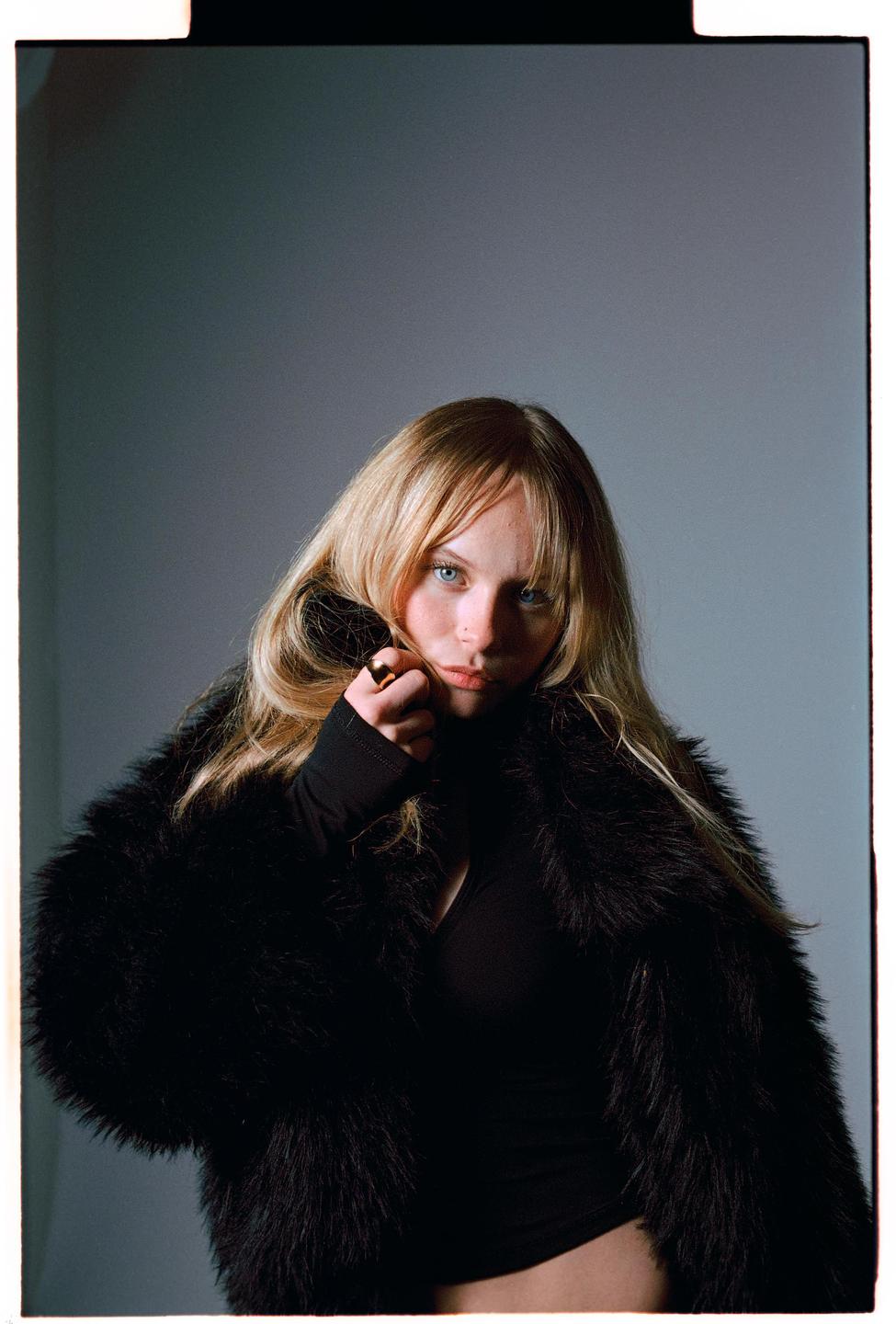 A woman with long blonde hair and blue eyes wears a black fur coat and a black top.