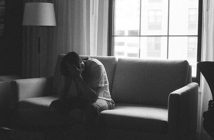 A man sits on a sofa with his face in his hands, next to a window with a city view.