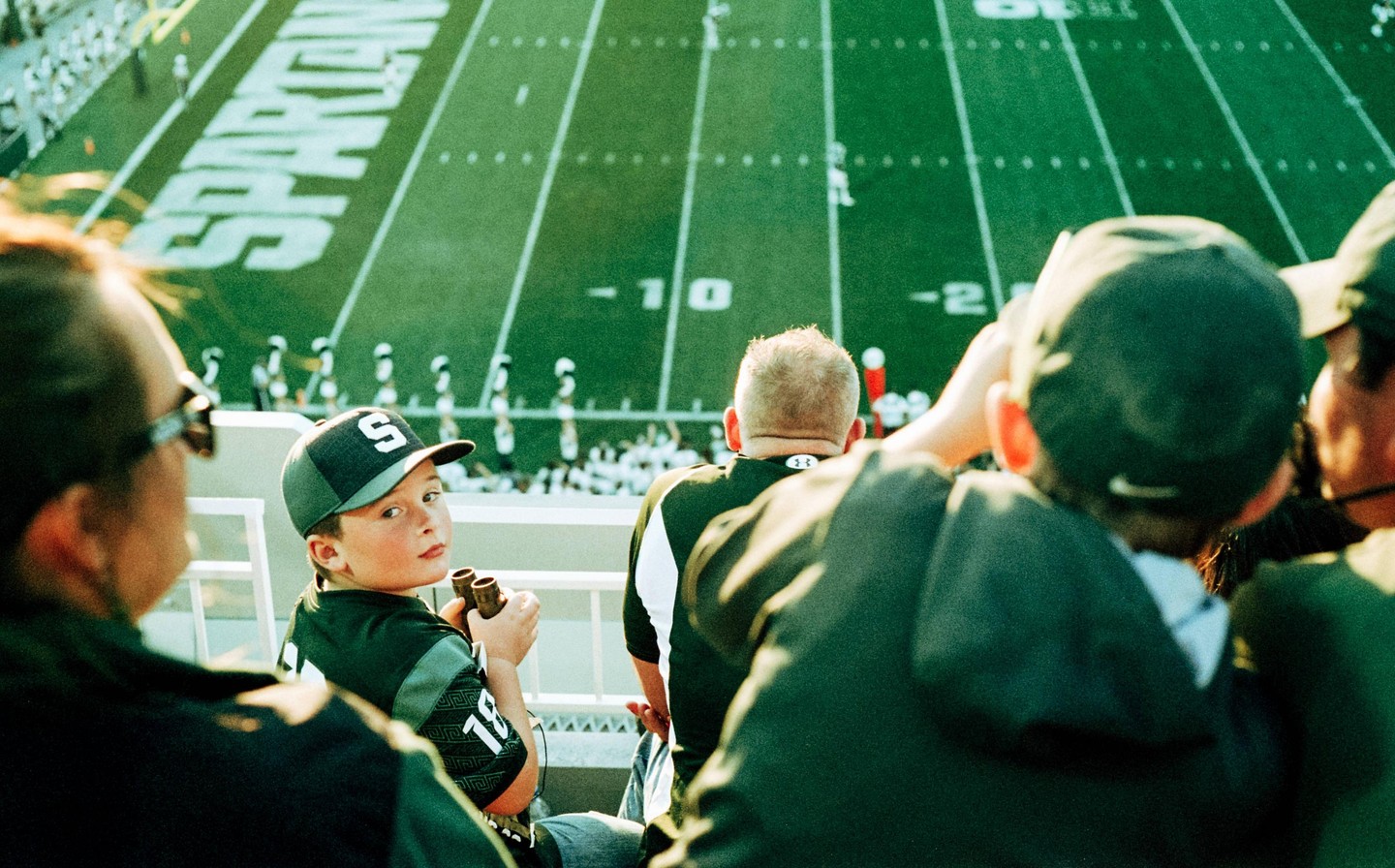 A boy in a baseball cap looks back at the viewer from a stadium seat overlooking a football field.