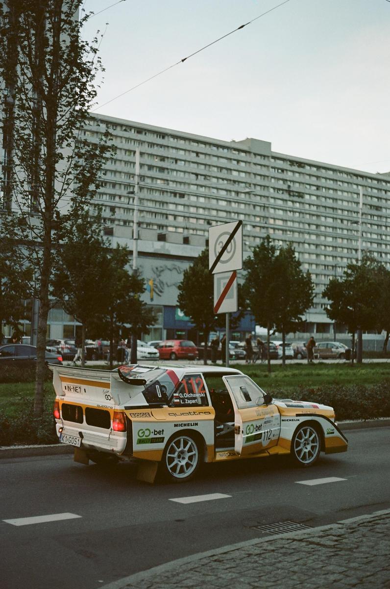 A white and yellow rally car with an open door is parked on a street next to a tall building.