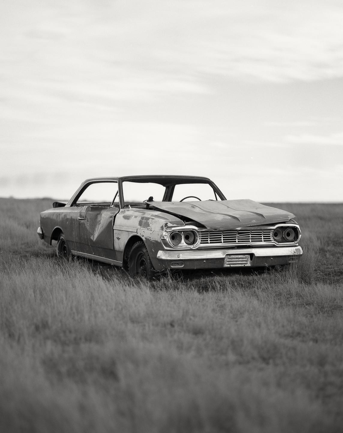 A wrecked car sits in a field of tall grass under a cloudy sky.