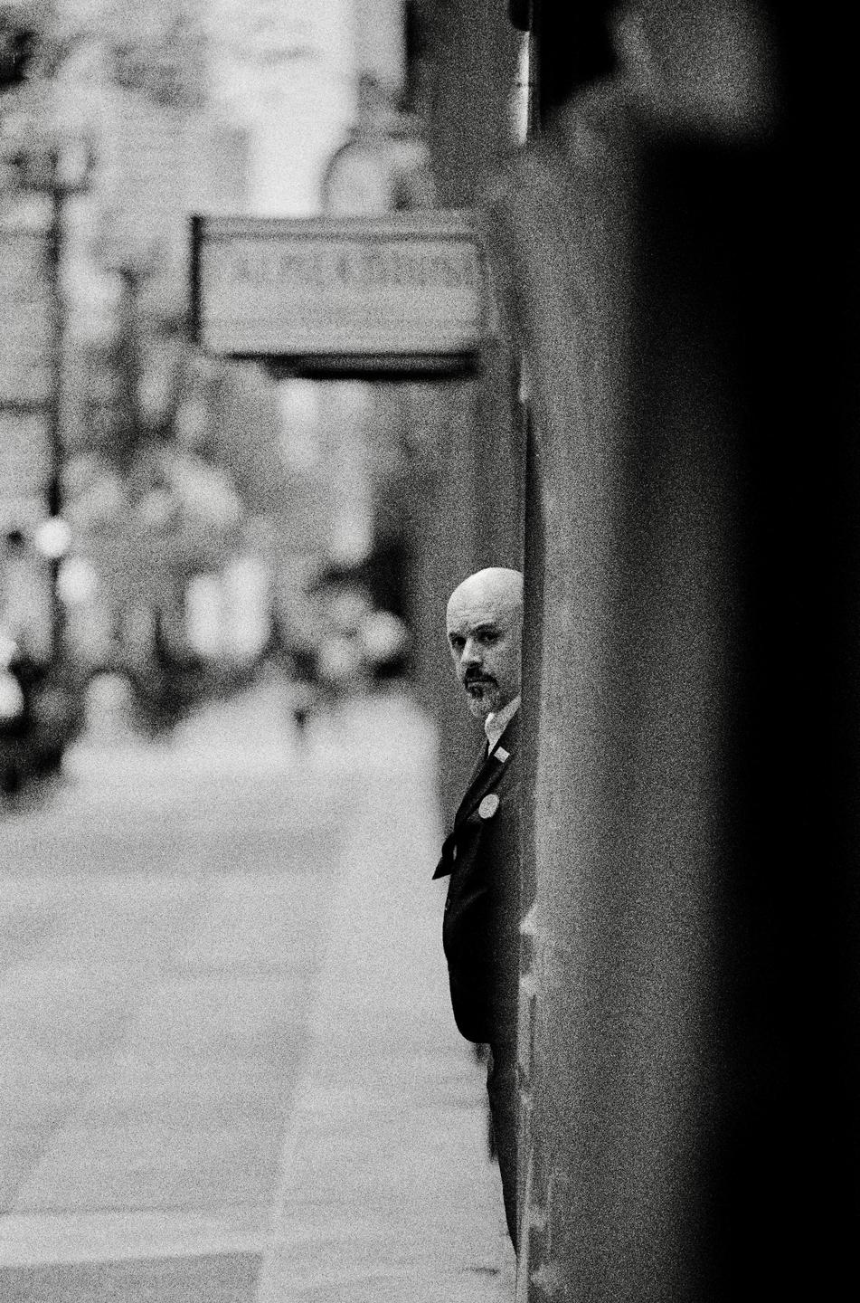 A man with a bald head and goatee looks out from behind a wall on a street.