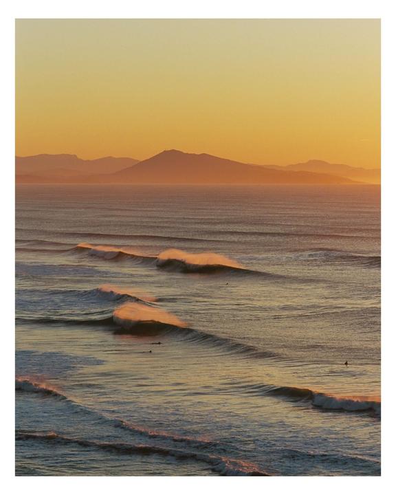 Waves roll towards the shore with mountains in the distance under an orange sky.