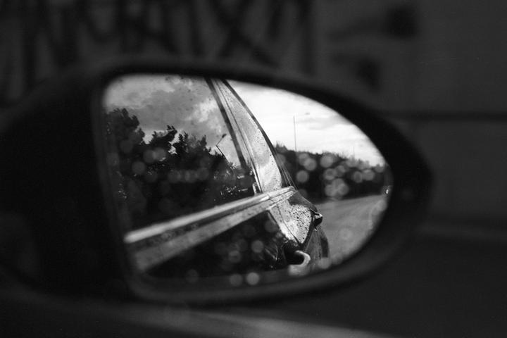 A car's side mirror reflects a road, trees, and sky, with raindrops on the mirror's surface.