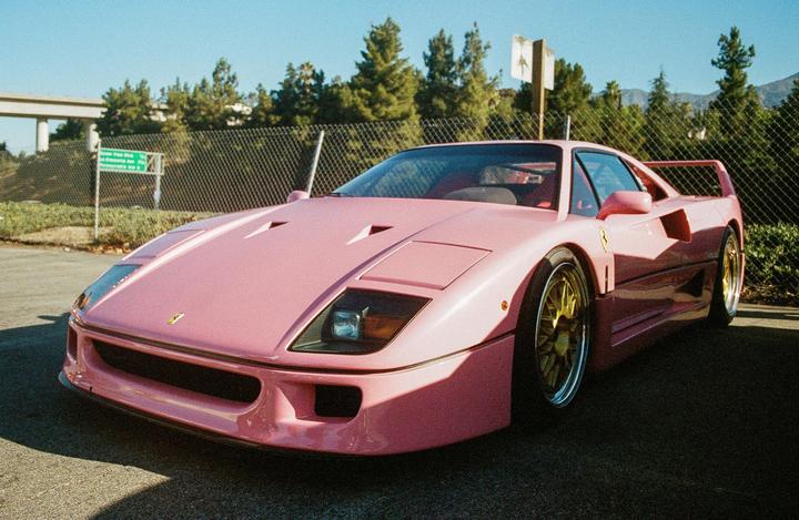A pink sports car with gold wheels is parked on asphalt next to a chain-link fence and trees.