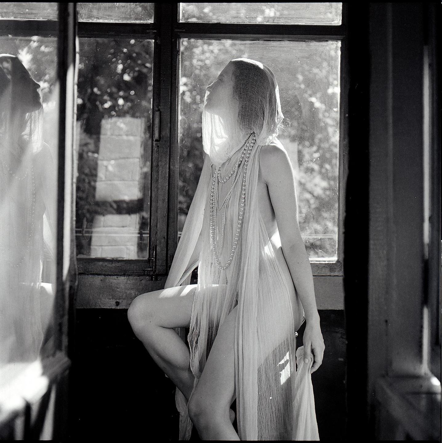 A woman draped in sheer fabric and pearls sits by a window, looking upwards.