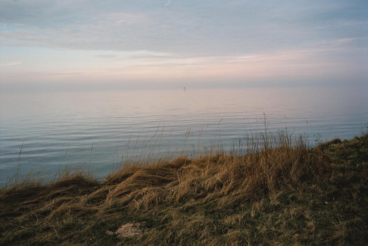 Dry grass grows on a hill overlooking a calm body of water under a hazy sky.