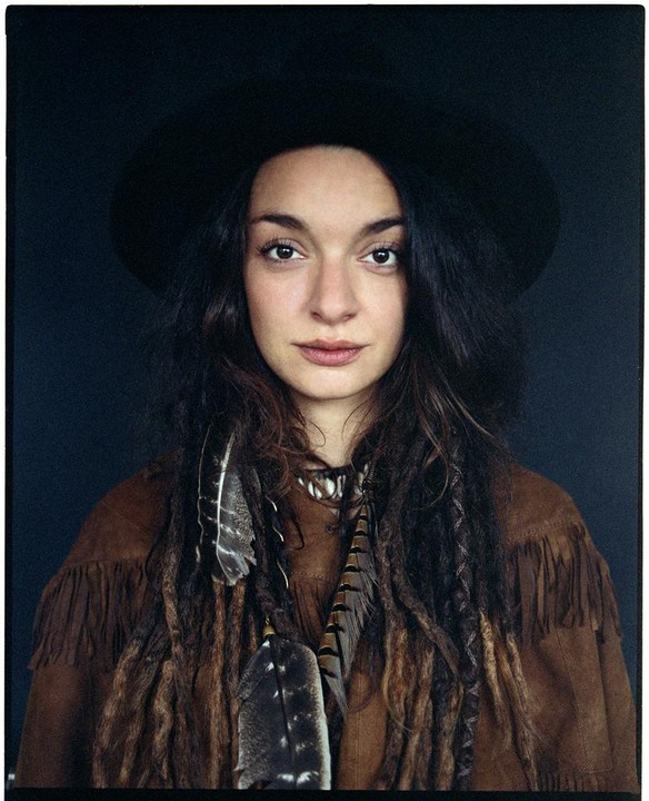 A woman with long, dark hair and a hat wears a fringed jacket and feather adornments.